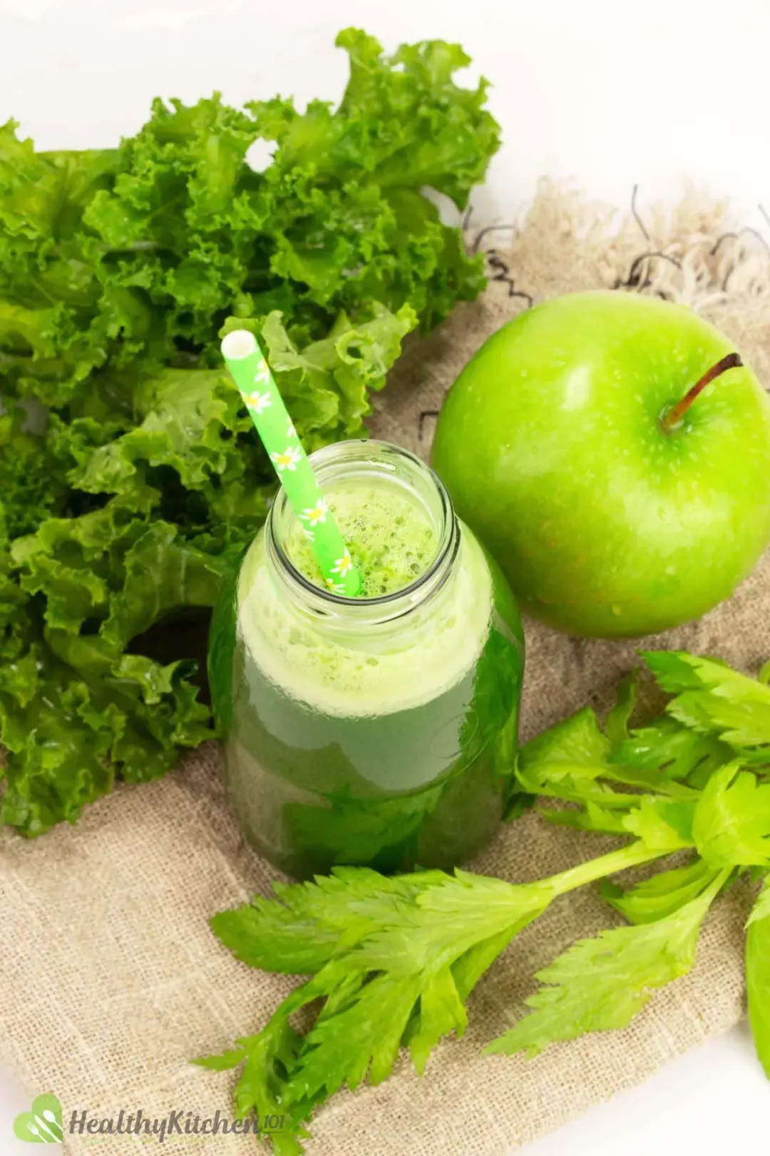 A short bottle of green vegetable juice with a green straw placed on a piece of fabric along with celery leaves, kale, and a whole green apple 