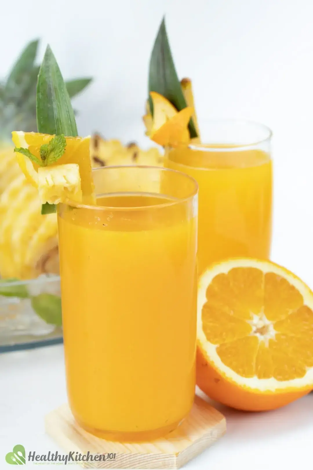 Two glases of orange pineapple juice garnished with a pineapple leaf, orange wedge, pinepple piece, next to half an orange