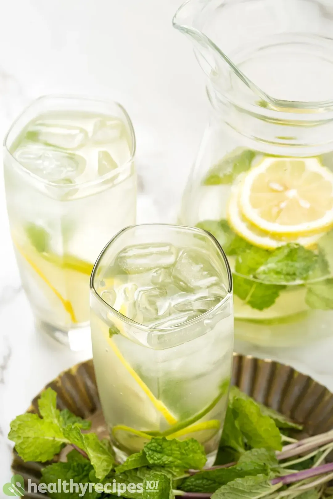 Two glasses and a pitcher of lemon water and mints, next to lots of mint leaves