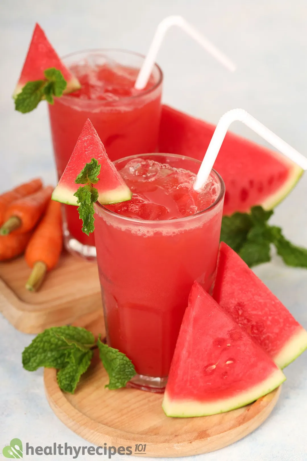Two glasses of watermelon carrot juice placed on wooden boards near mint leaves, watermelon slices, and carrots.