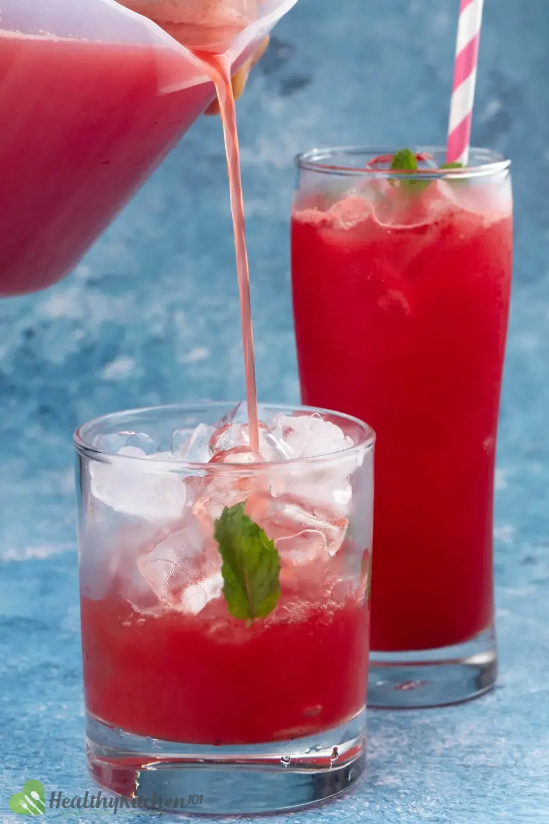 Watermelon juice being poured into a glass filled with ice and a mint leave, there’s a tall glass of watermelon juice in the back