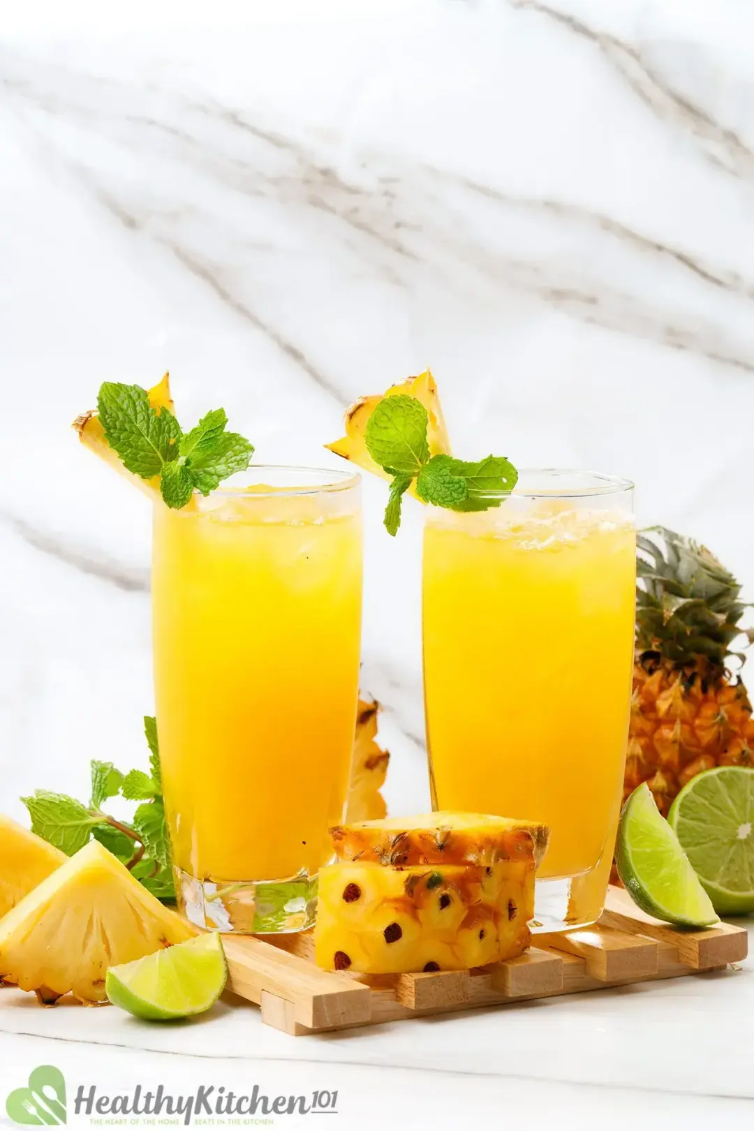 Two identical glasses of pineapple juice and cider cocktail with mints on the rim and next to pineapple wedges, lime wedges