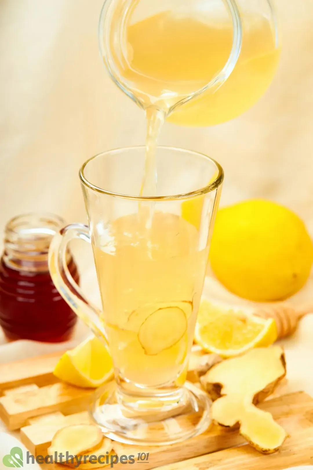 A pitcher of lemon honey ginger vinegar drink pouring into a shot glass with handle, next to some lemons and ginger