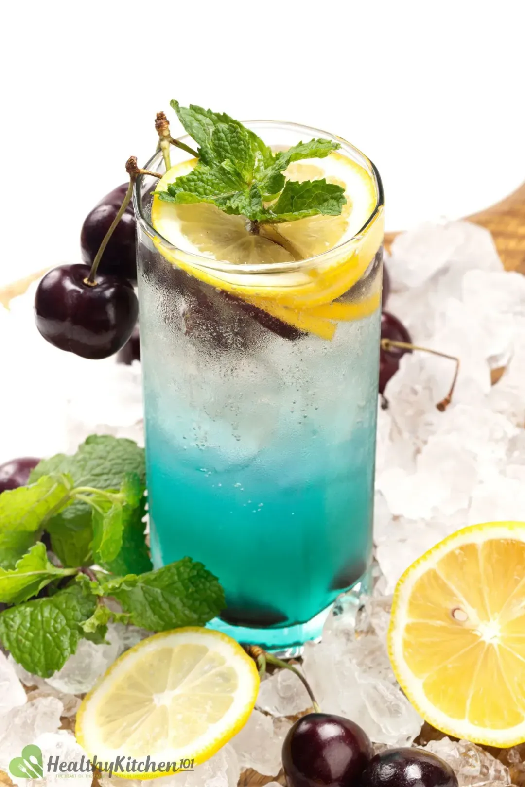 A cocktail glass of a cyan-hued drink, topped with cherries and lemon wedges, with lemon wheels and mints lying around