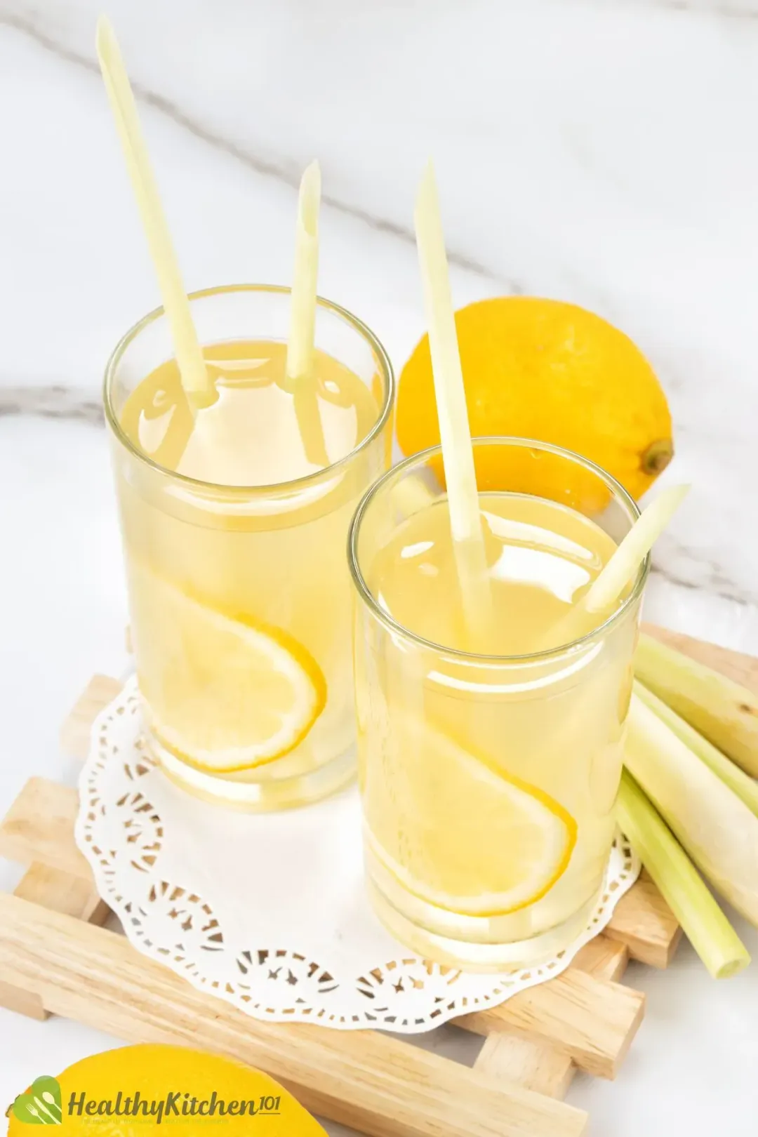 Two glasses of lemon juice and cider drinks with lemon wheels inside, next to whole lemons and lemongrass stalks