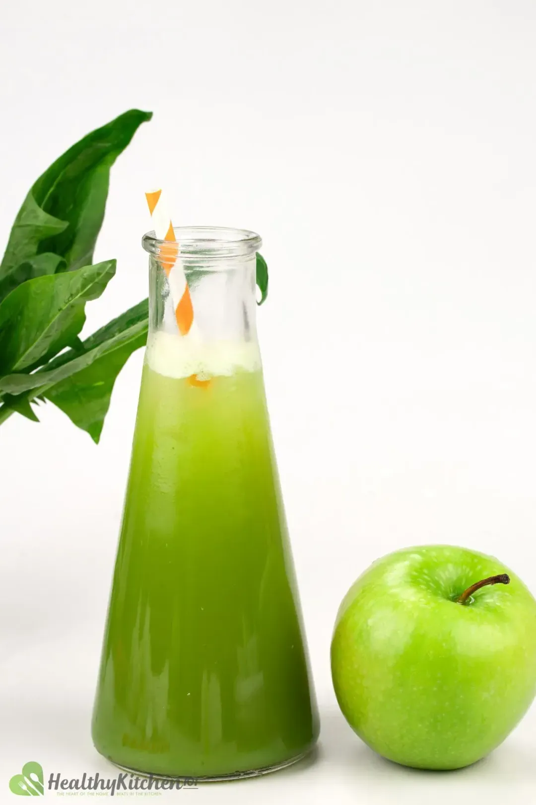 A pitcher of green apple juice decorated with an orange-striped straw alongside a whole green apple and spinach