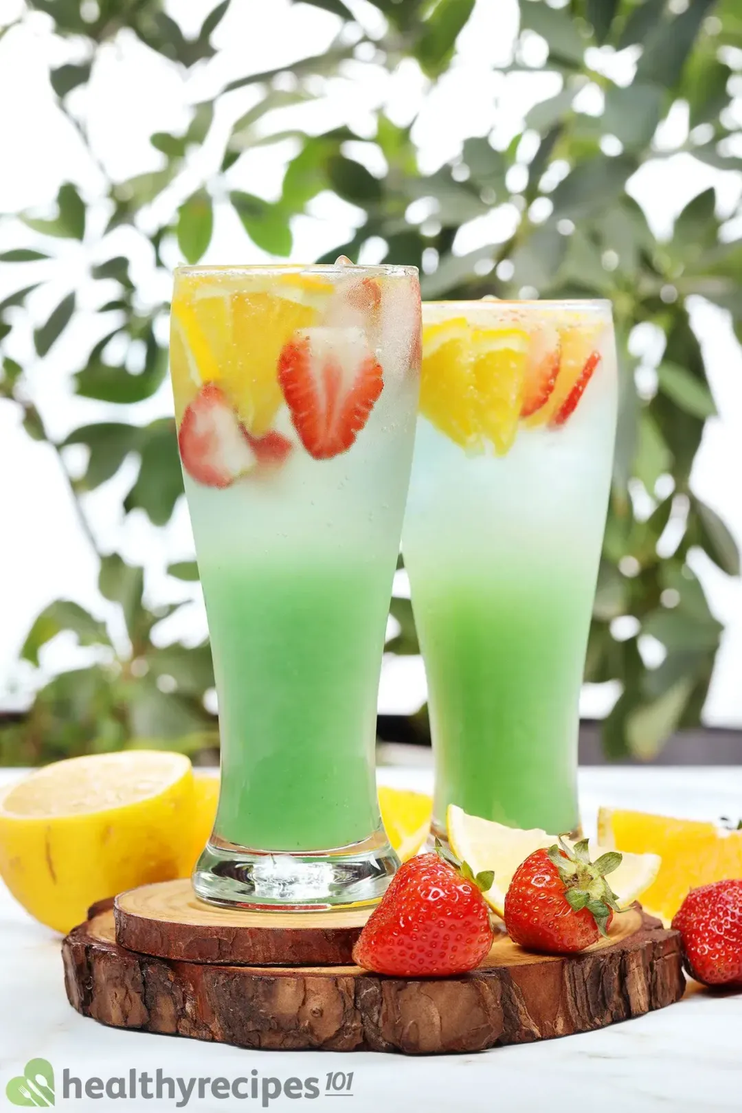 In front of a tree, two glasses of a green jungle juice cocktail put next to whole strawberries and whole lemons
