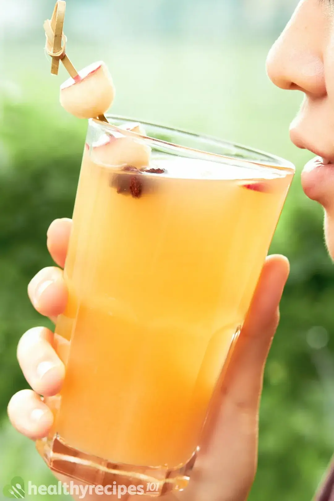 A person holding a tall glass filled with apple juice garnished with an apple balls skewer