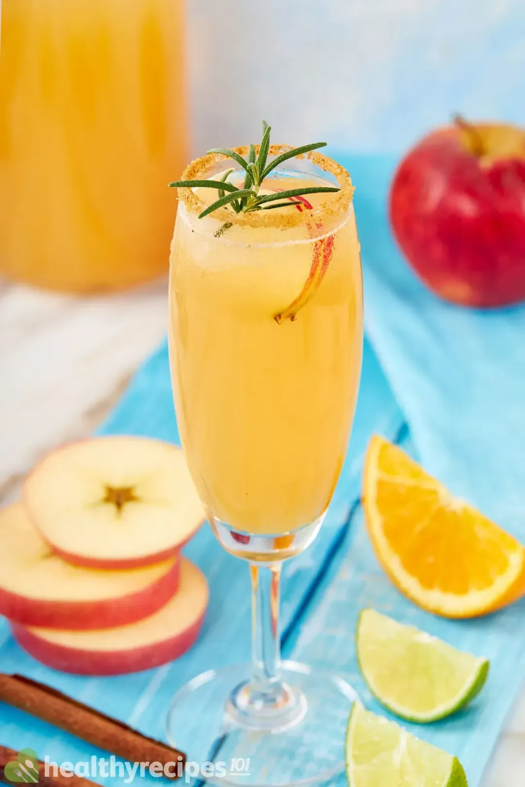 A cocktail glass of apple juice margarita garnished with rosemary, apple slices, and citrus wedges on a blue table