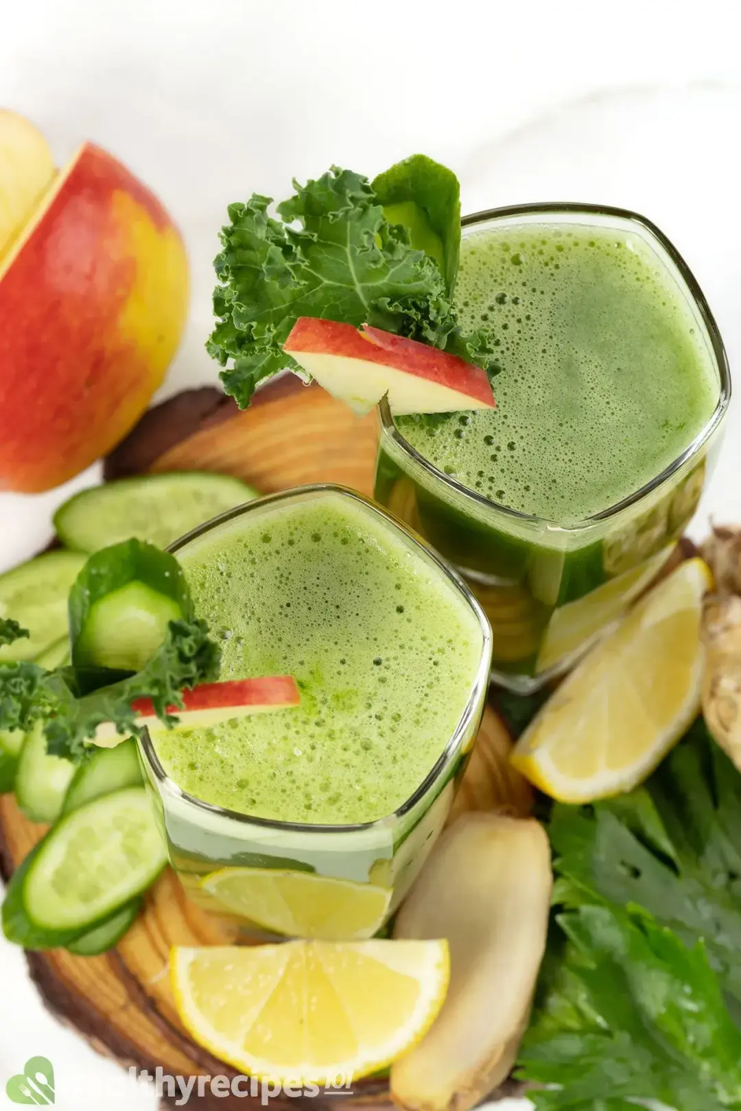 Two glasses of green juice garnished with lemon wedges, cucumber slices, and kale, taken from above