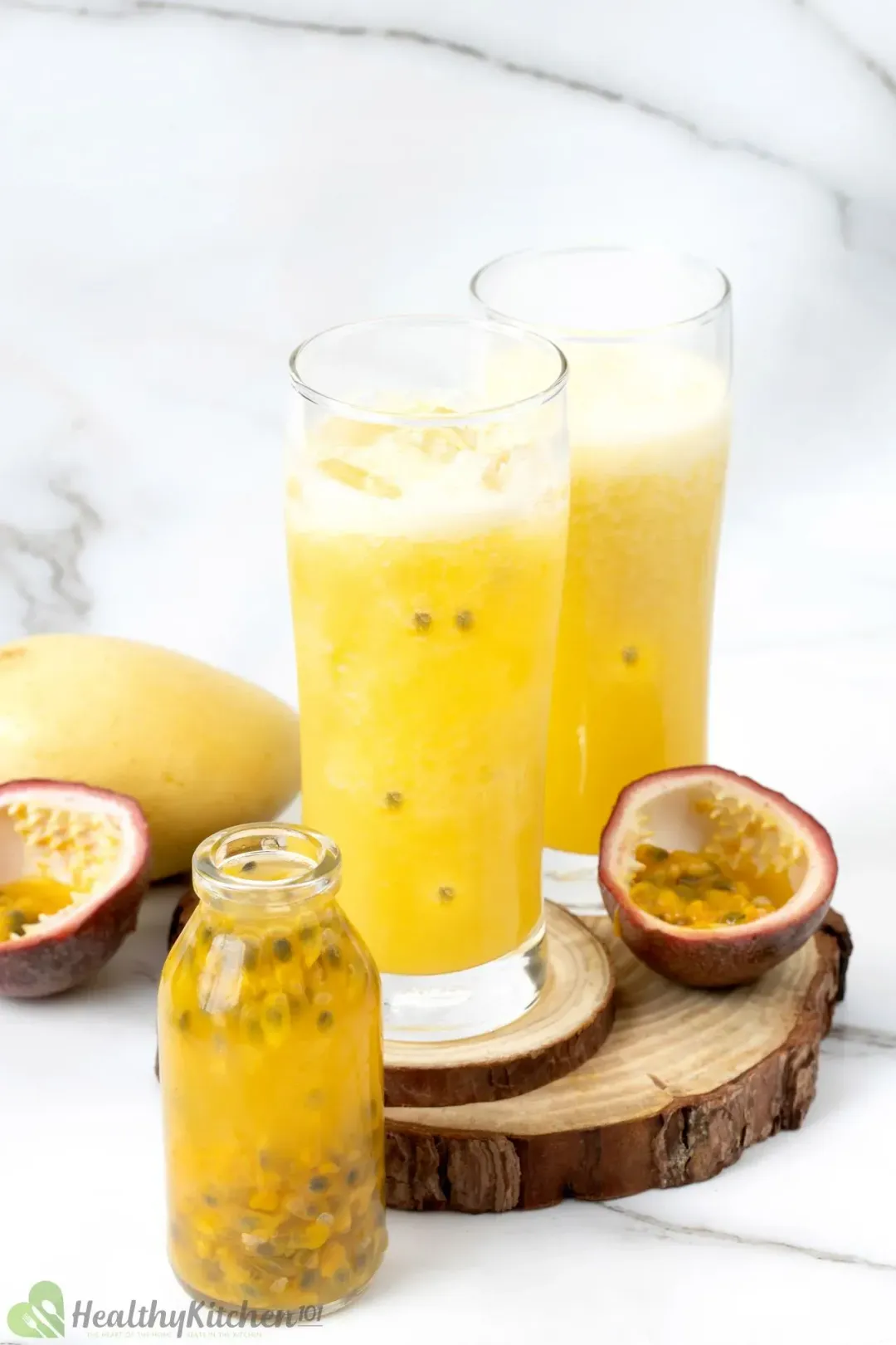 Two glasses of passion fruit juice on a wooden coaster with half a passion fruit, next to a jar of passion fruit juice