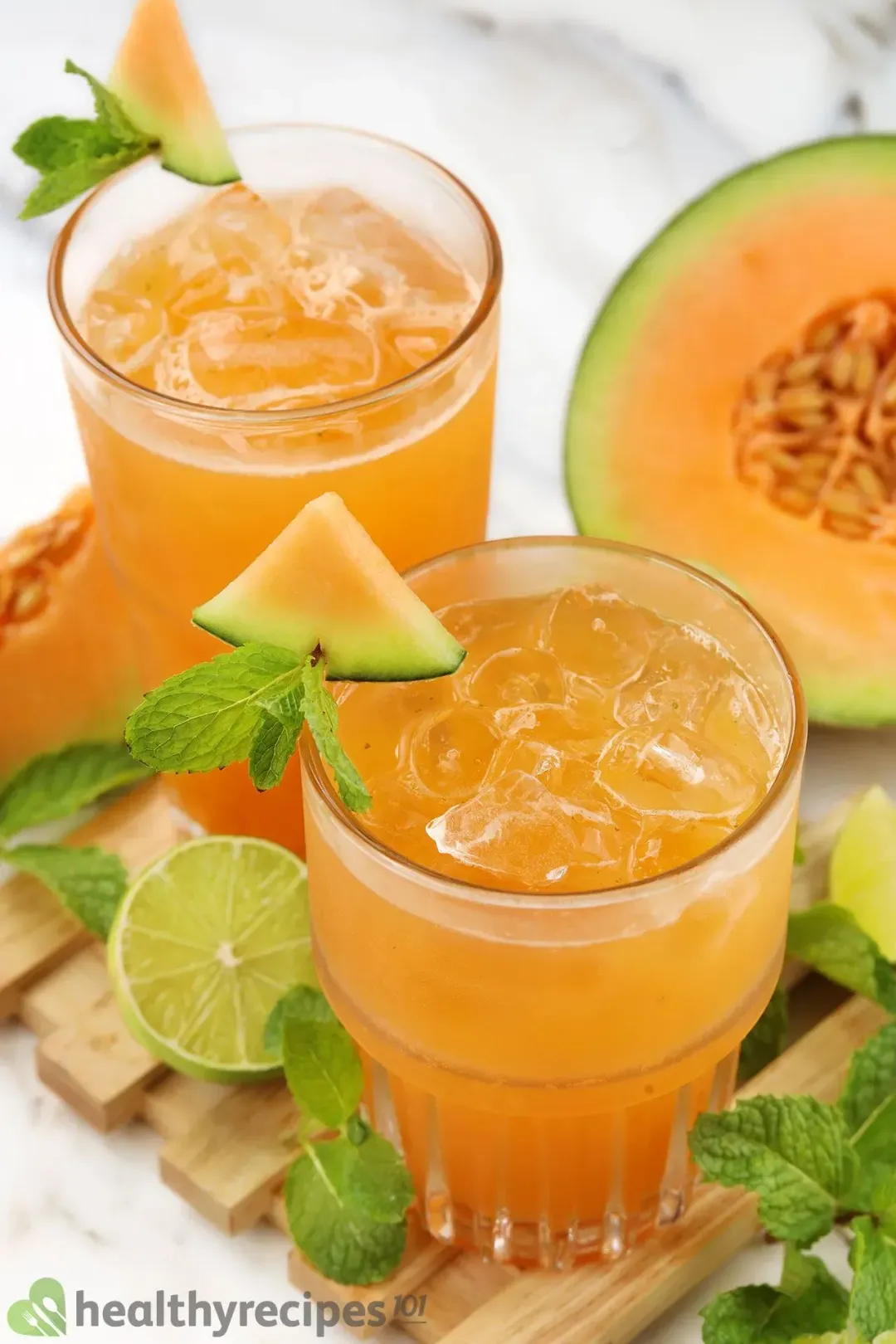 Two iced glasses of cantaloupe drinks, garnished with mints, cantaloupe wedges, and half of a lime