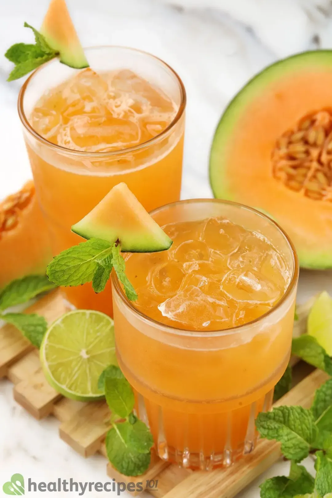 Two iced glasses of cantaloupe drinks, garnished with mints, cantaloupe wedges, put next to half of a cantaloupe