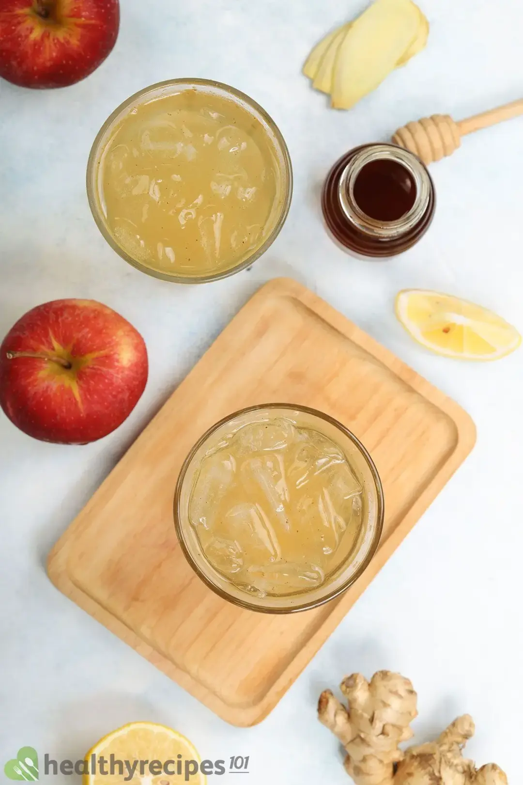 A shot taken from above: two iced glases of apple cider vinegar drinks on wooden tray next to apples, pineapples, ginger, and a honey jar