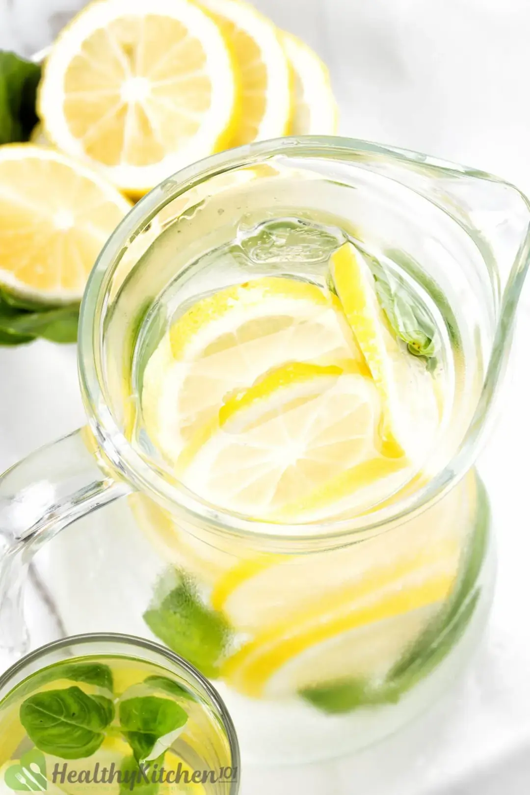 A close-up shot of a lemon water pitcher, next to a bowl of lemon wheels on basil leaves