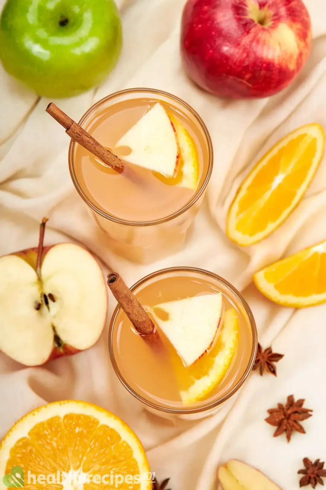 Two glasses of homemade apple cider with cinnamon sticks, apple triangles, orange triangles, and whole apples around