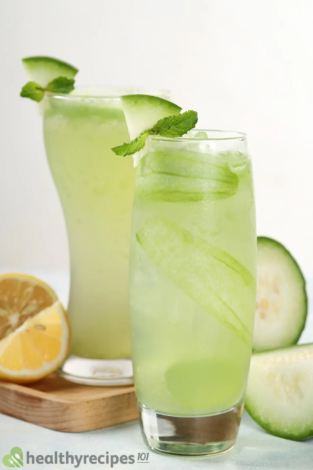 Two tall glasses of winter melon juice, one of which is placed on a wooden board near lime wedges and slices of winter melon.