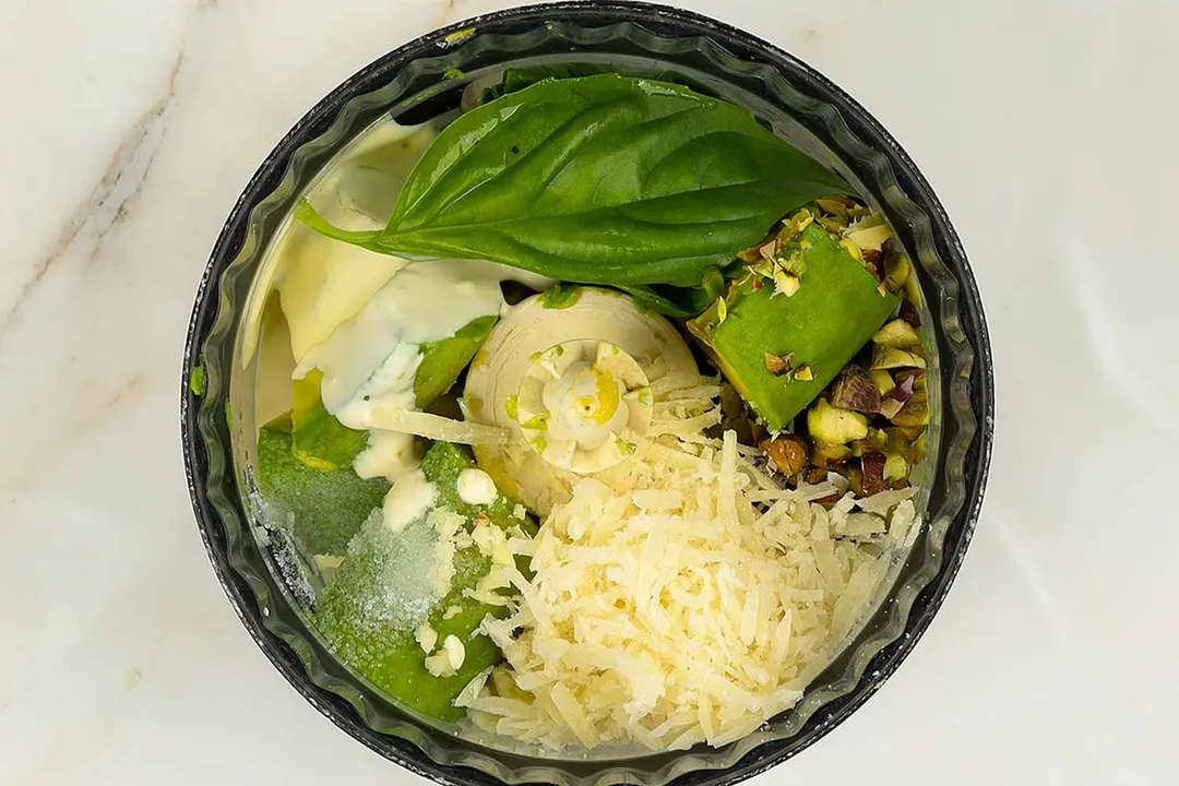 A food processor filled with grated cheese, leafy greens, nuts, and avocado slices.