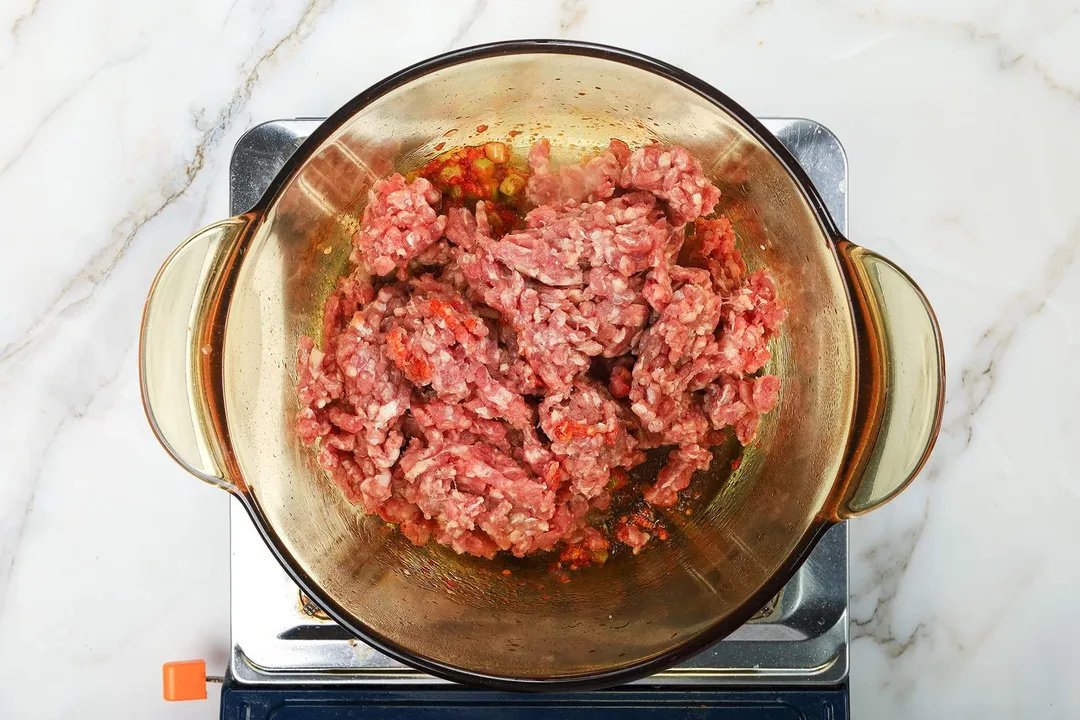 A saucepan cooking raw ground beef on a portable gas stove.