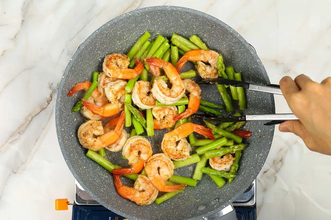 A hand using a pair of tongs to toss cooked shrimp and asparagus cooking on a pan.