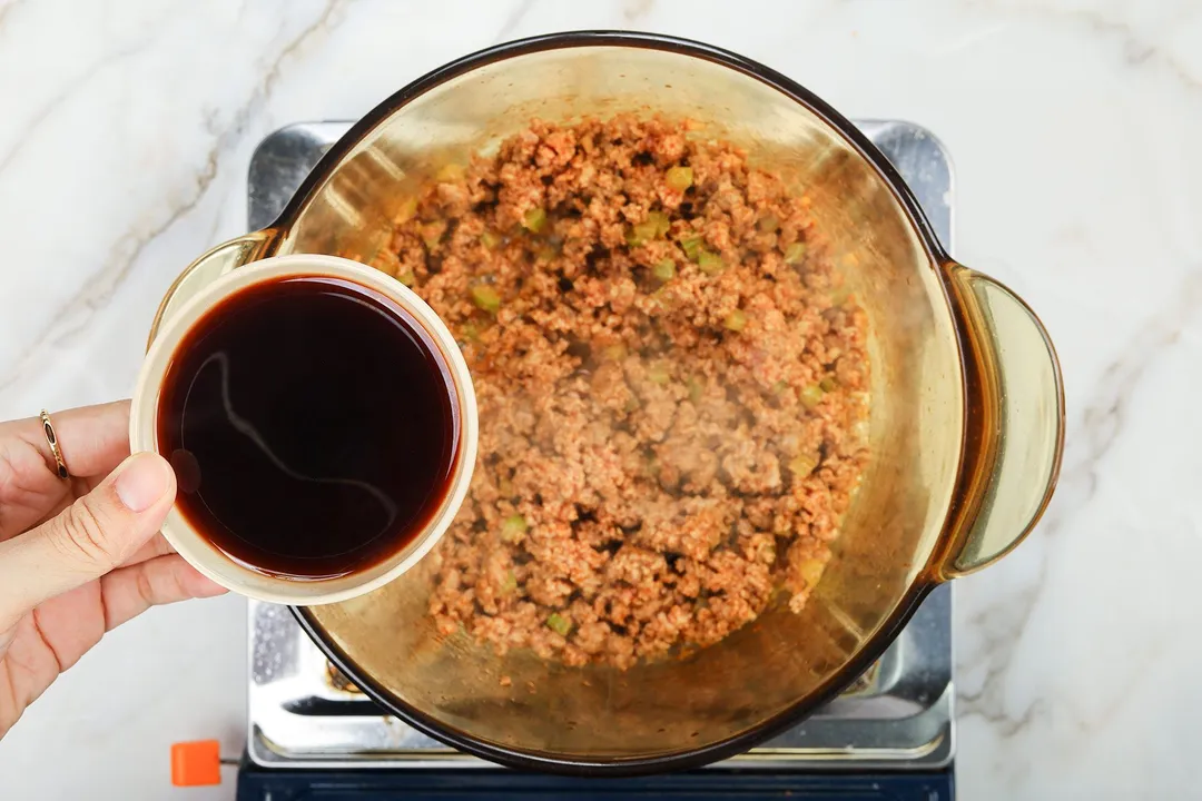 A hand holding a small bowl of red wine over a sauce pan cooking ground beef.
