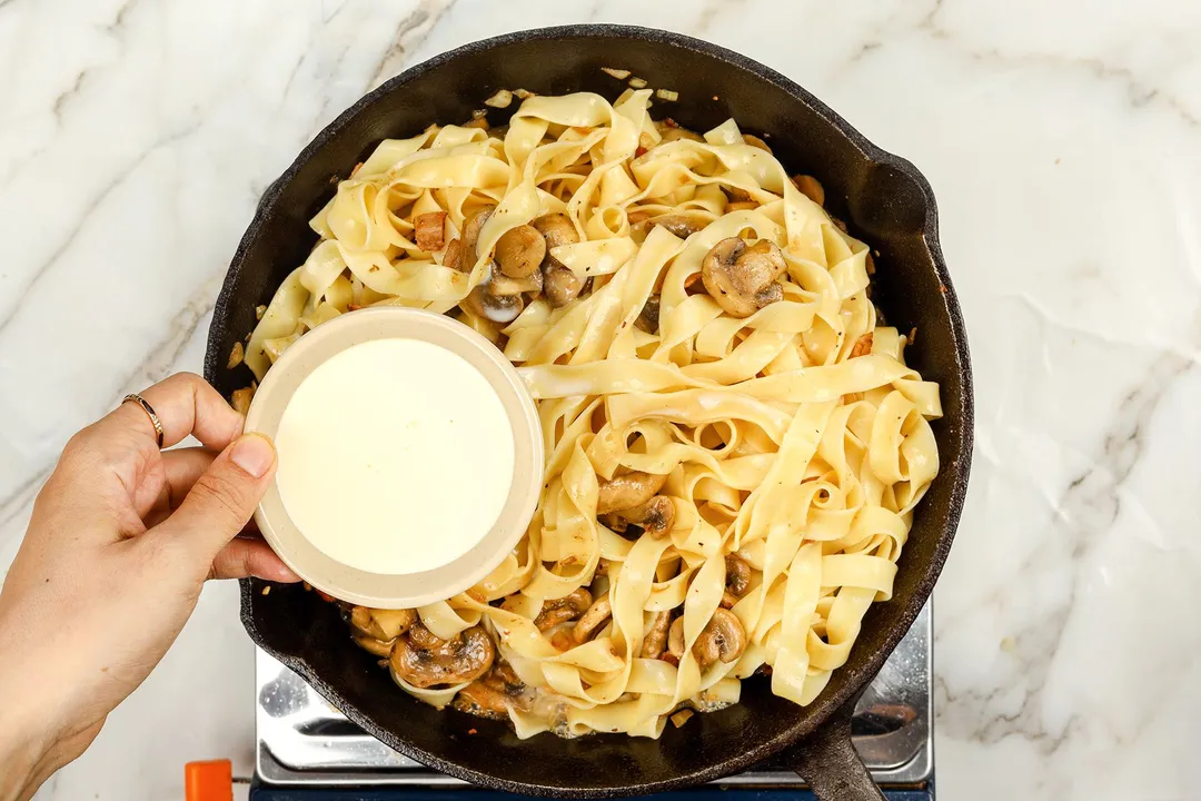 hand holds a small bowl of heavy cream on top of a skillet of pasta