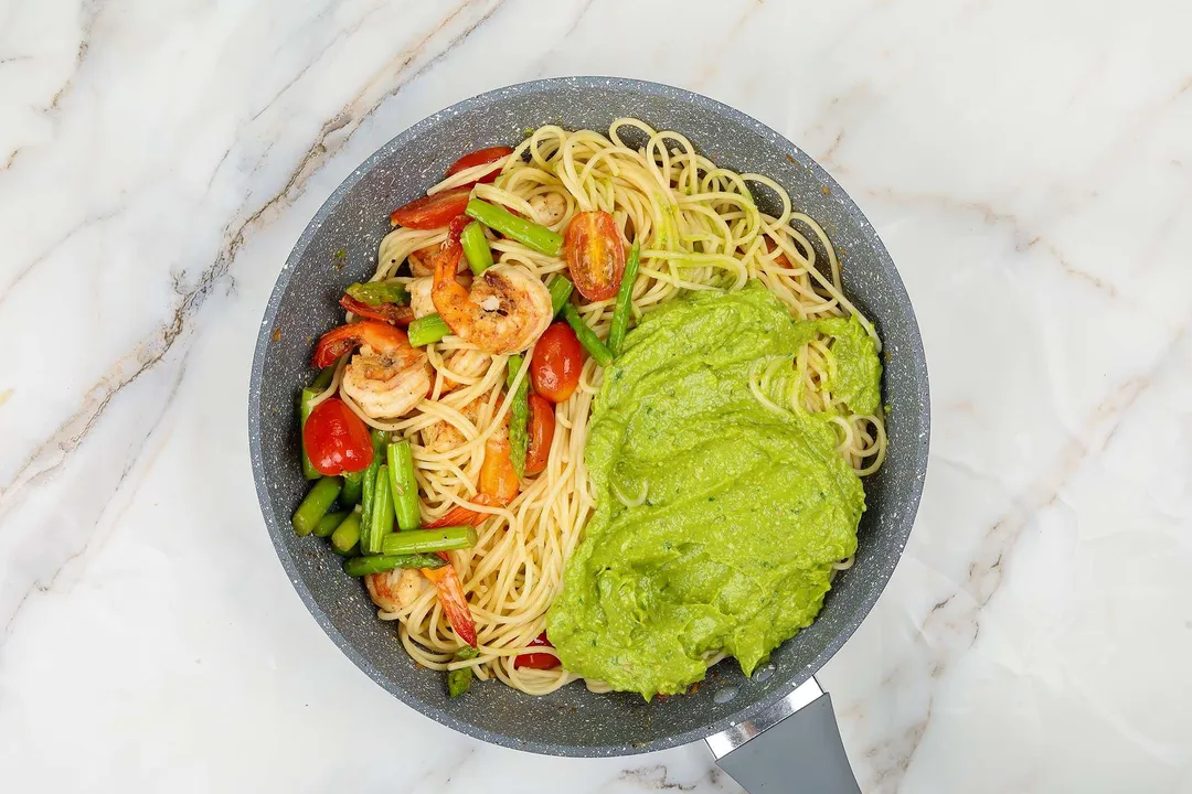 A pan cooking spaghetti, shrimp, and vegetables covered in a creamy green avocado sauce.