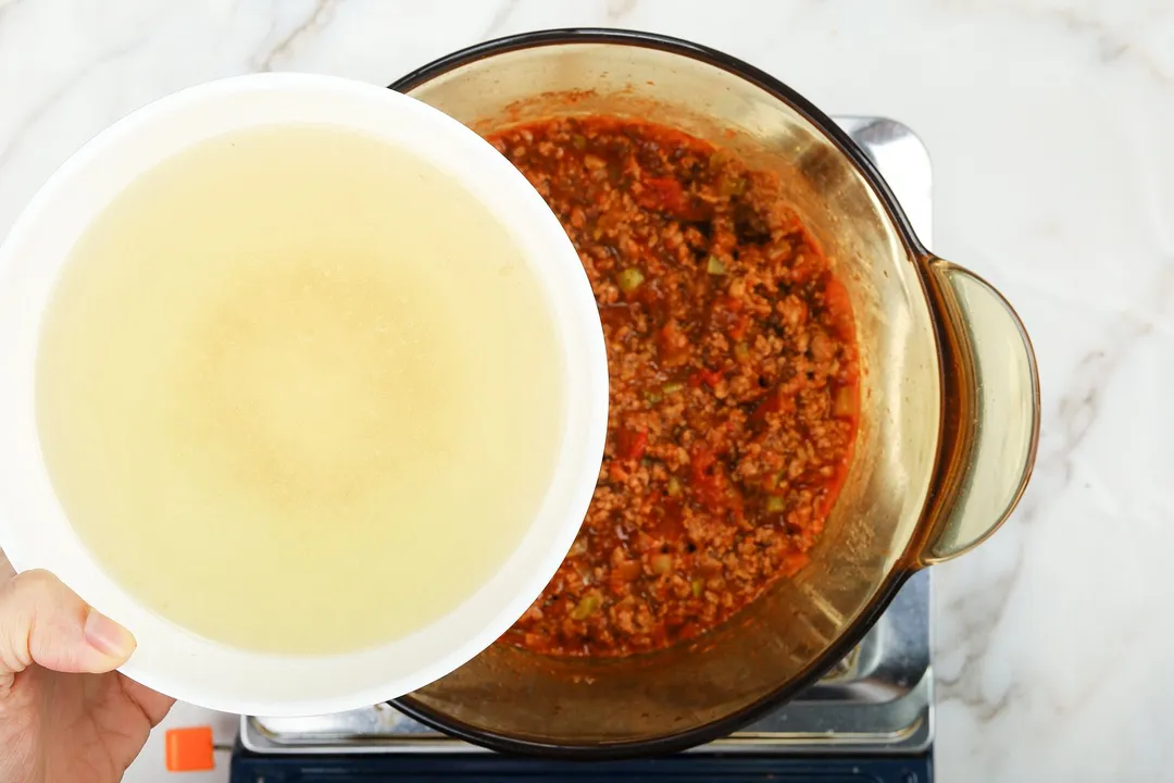 A handing holding a bowl of chicken broth over a saucepan cooking ground beef mixed with tomato sauce.