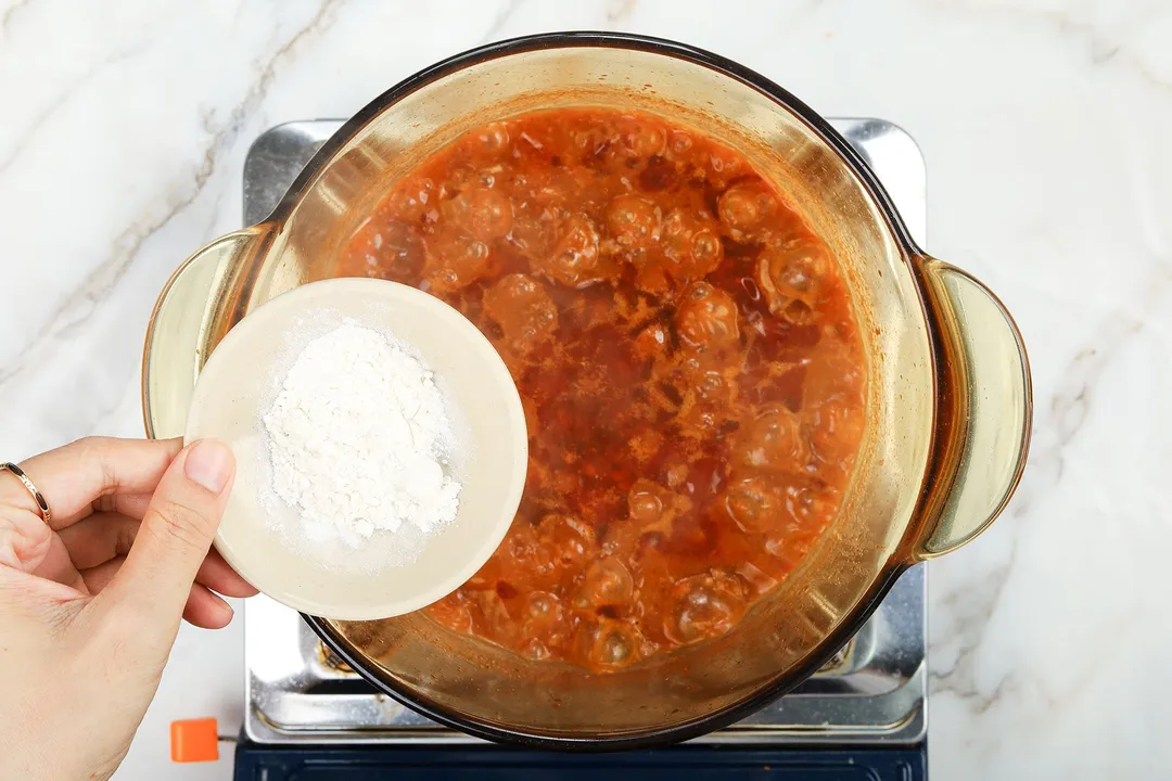 A hand holding a small plate of flour over a saucepan cooking ground beef mixed with tomato sauce.
