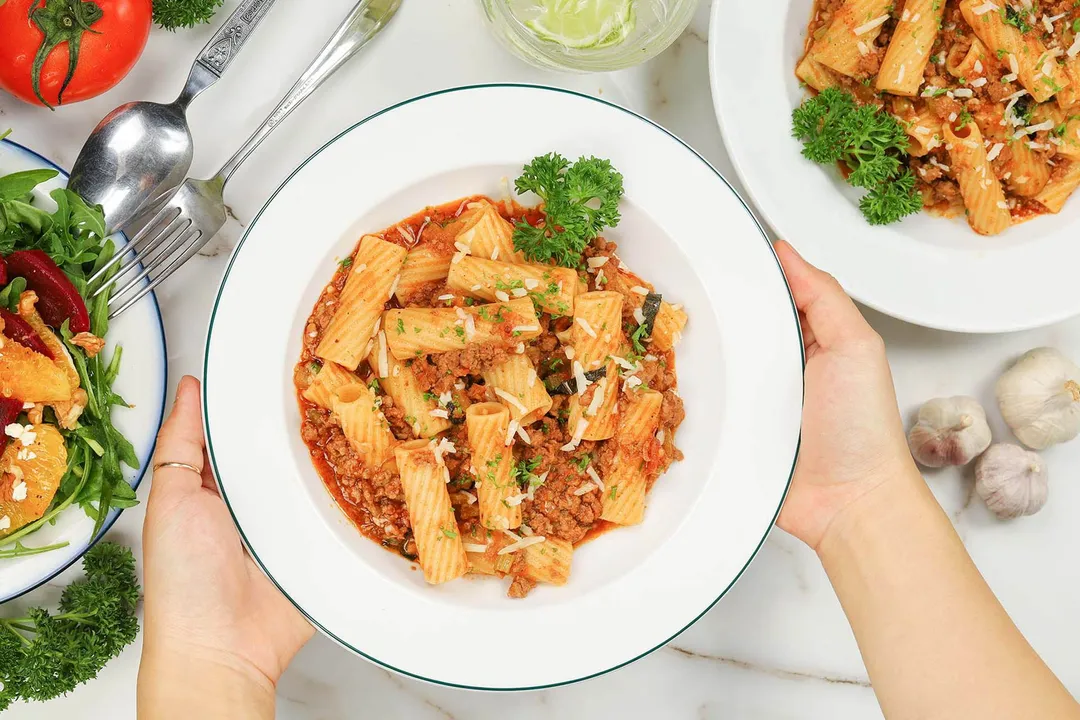 Two hands holding up a plate filled with tortiglioni pasta bolognese