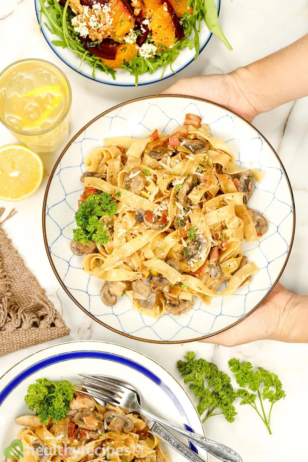two hand holds a plate of cooked mushroom pasta next to a plate of salad, a glass of juice
