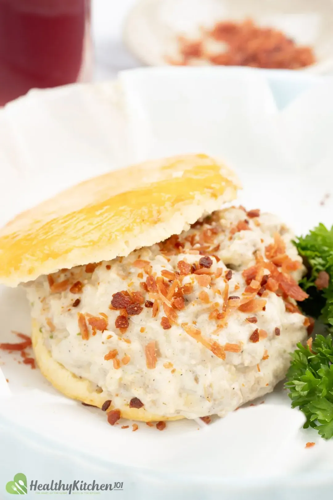 A heap of meaty gravy and bacon crumbles in between two biscuits, placed next to green parsley