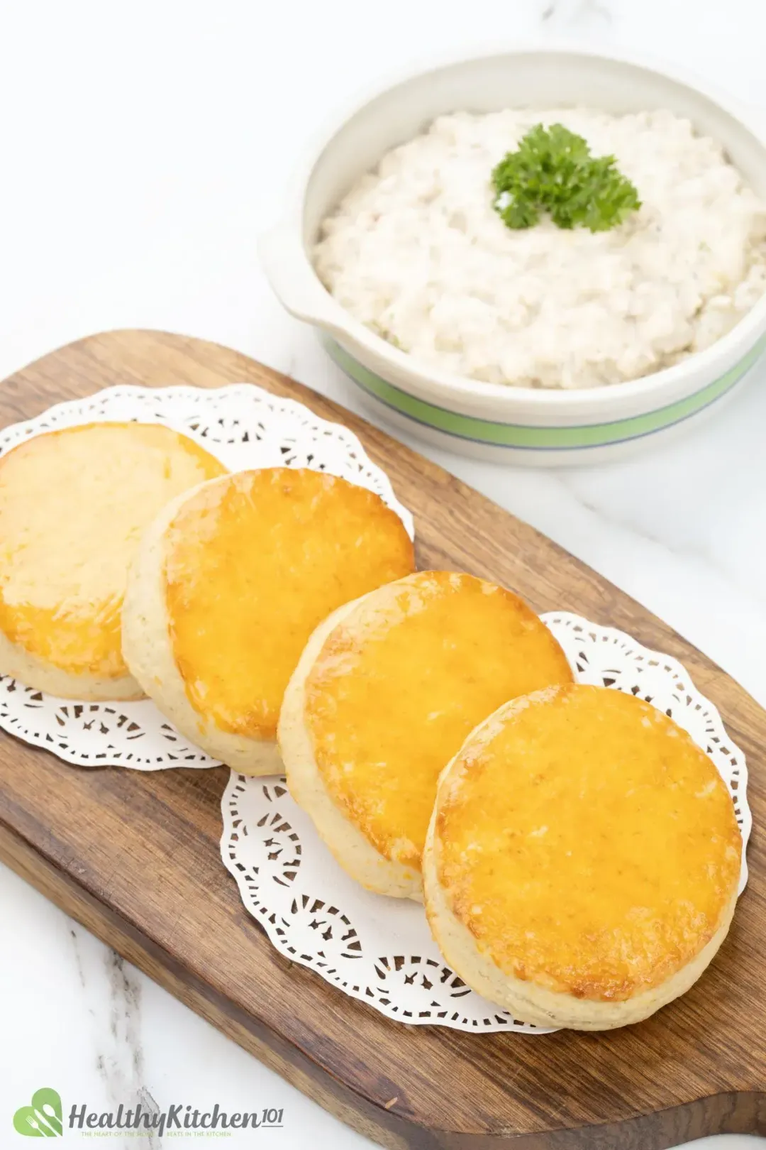 Four round biscuits on a wooden board next to a bowl of white gravy and parsley sprig