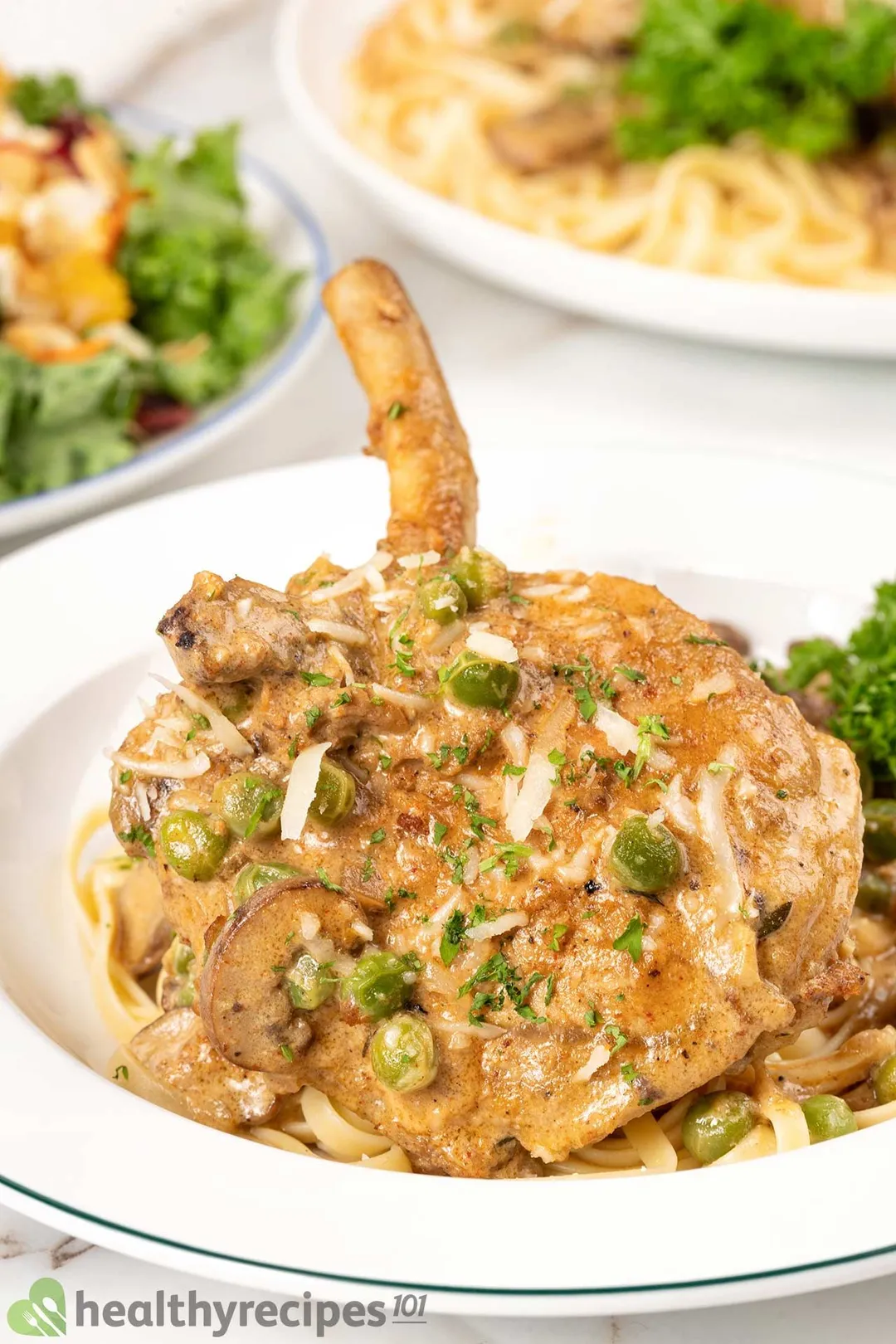 cooked pork chop with green beans, cheese and sauce bedding on pasta on a white plate with a salad and pasta plate in backgorund