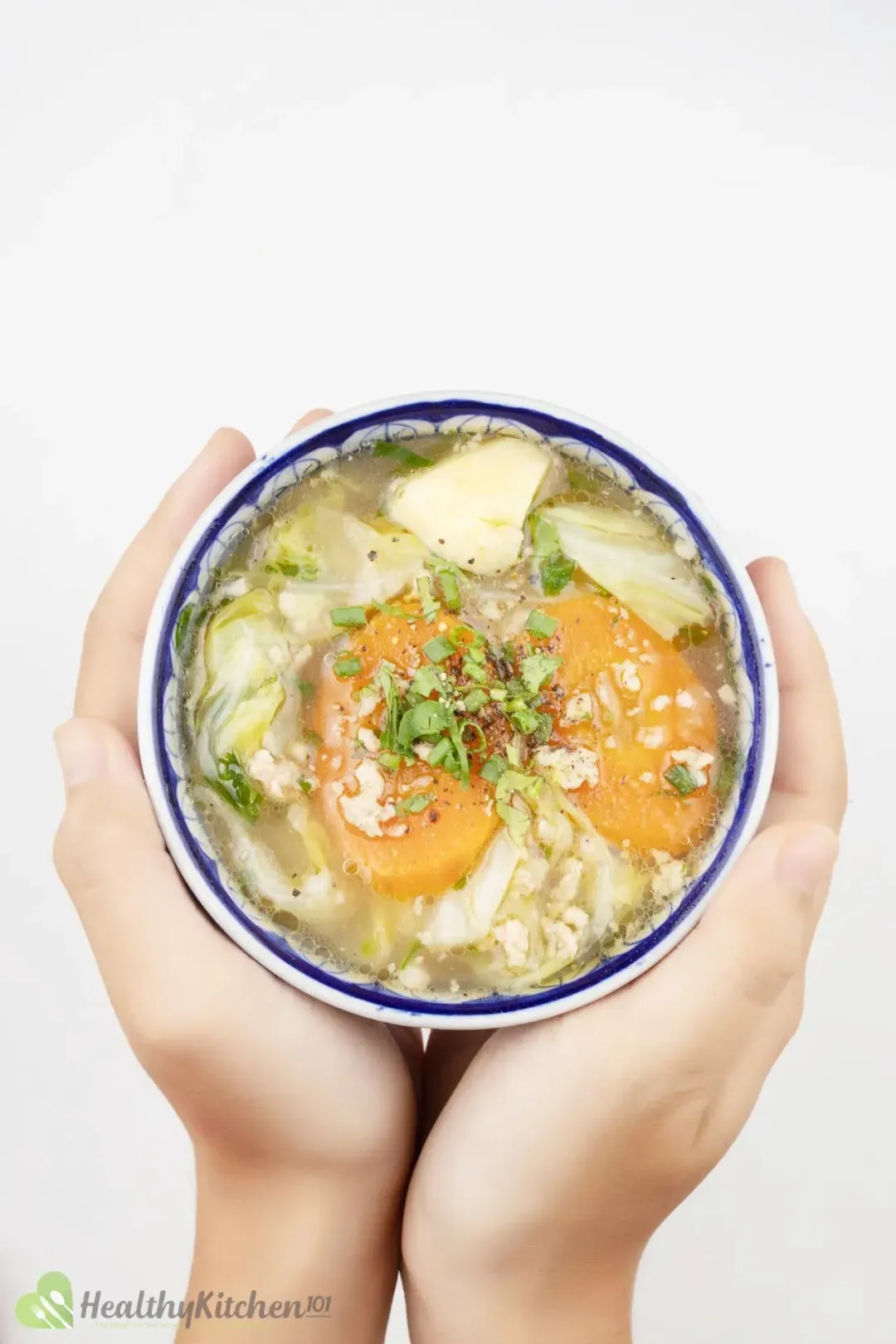 Two hands holding a Chinese porcelain bowl of cabbage, potato, carrots, ground pork, and green onions