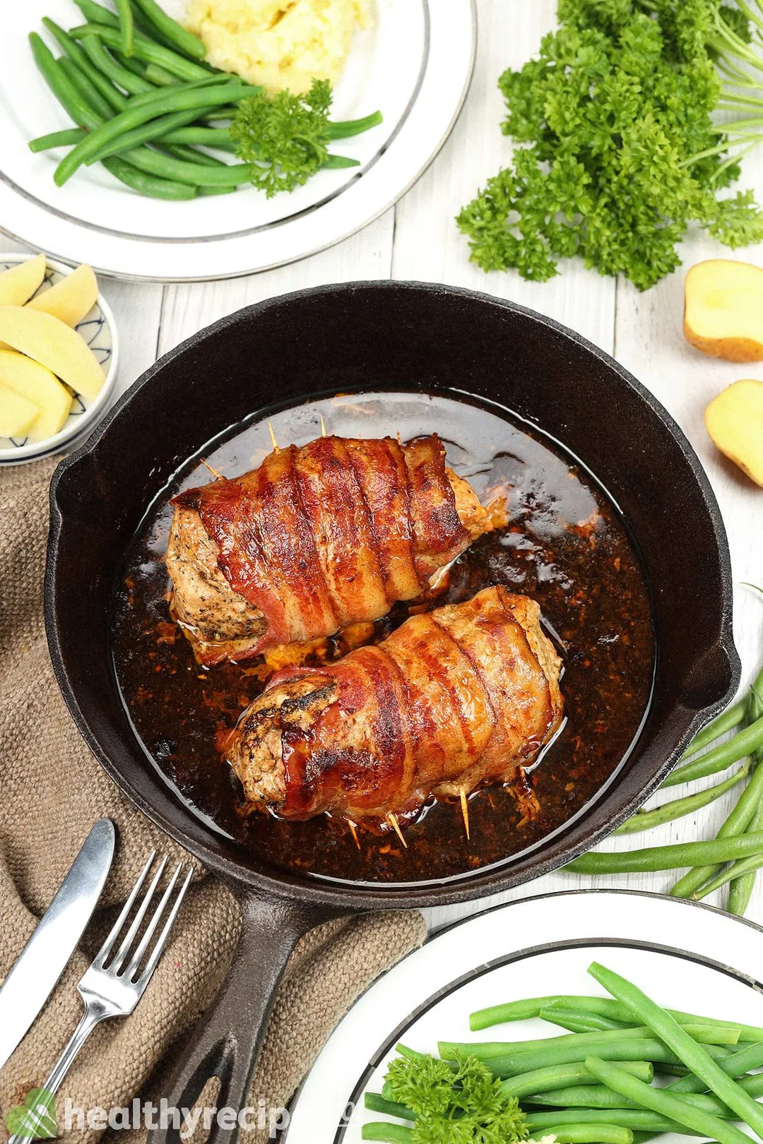 a cast iron skillet of two cooked bacon wrapped pork tenderloin decorated with green beans, potatoes