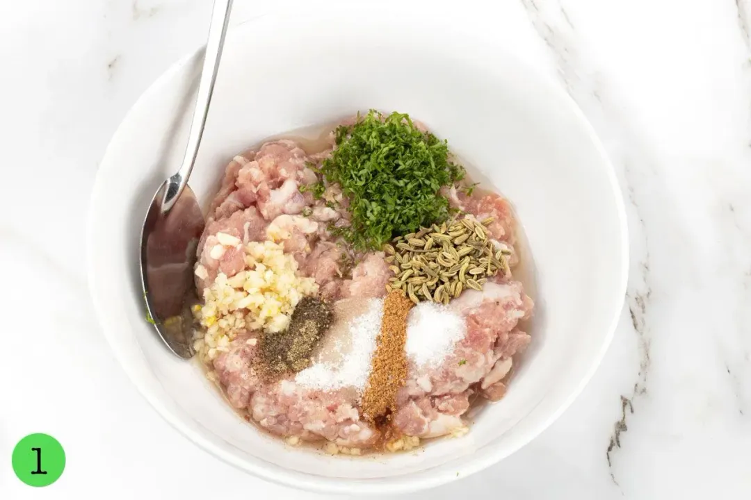 Ground meat, spices, and herbs in a white bowl, about to be mixed