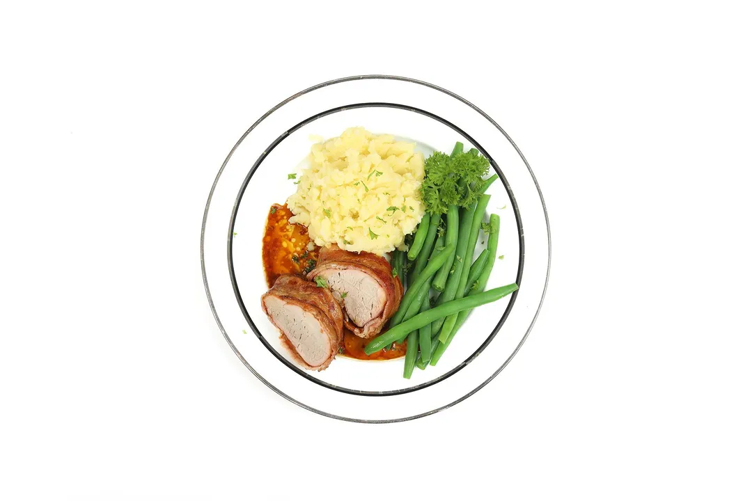 a plate of two slices of bacon wrapped pork tenderloin with mashed potato and green beans