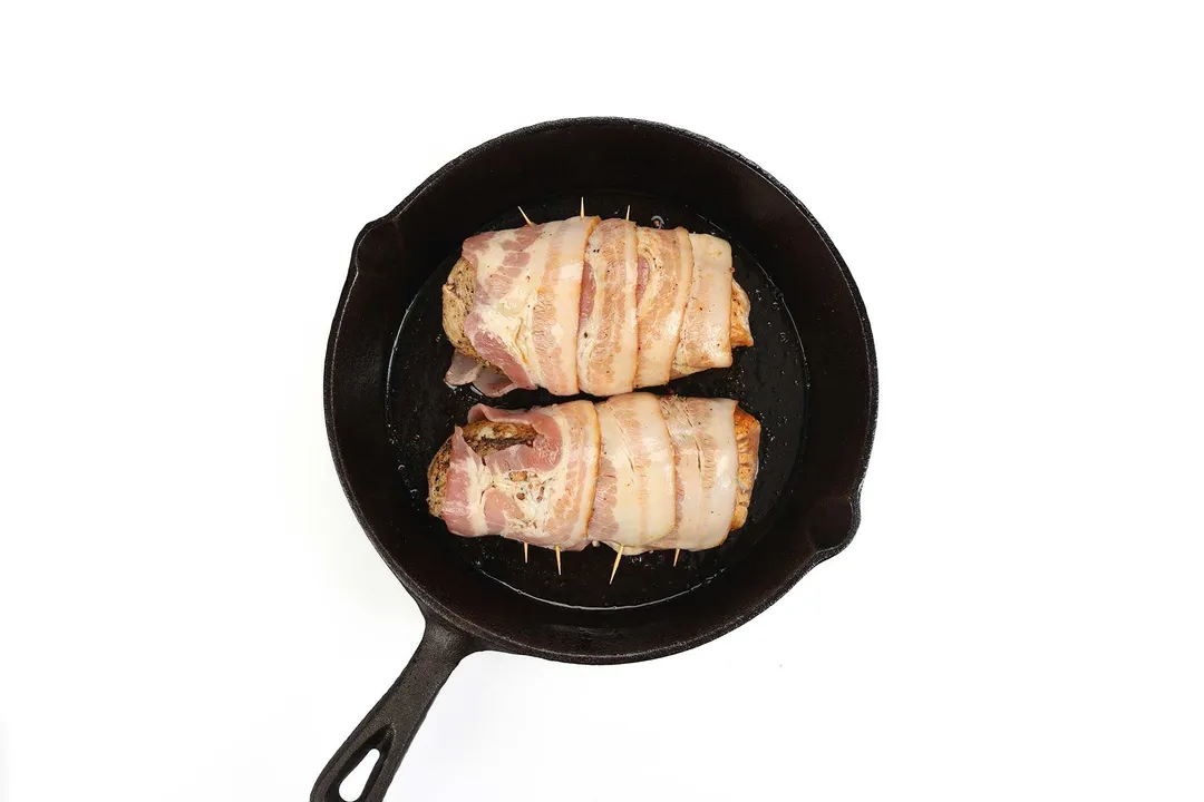 searing two bacon wrapped pork tenderloin in a cast iron skillet