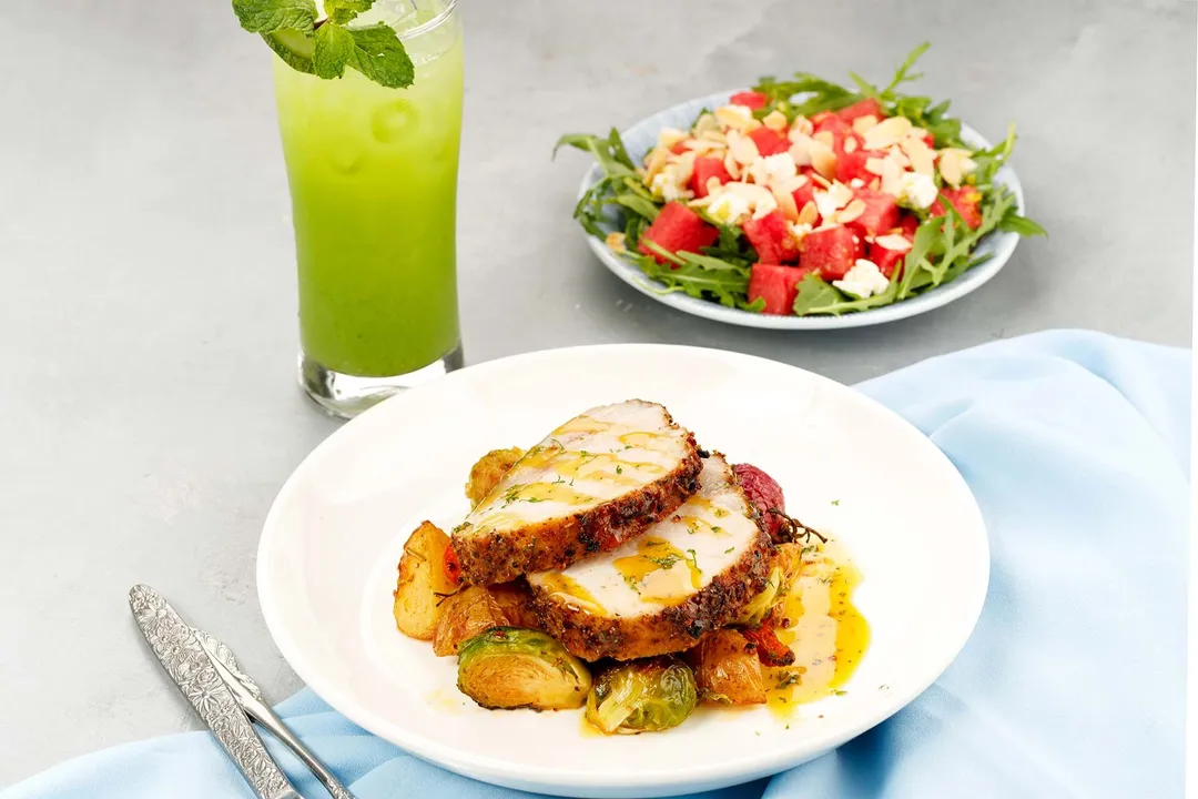 a plate of cooked pork loin next to a glass of green juice and a plate of salad