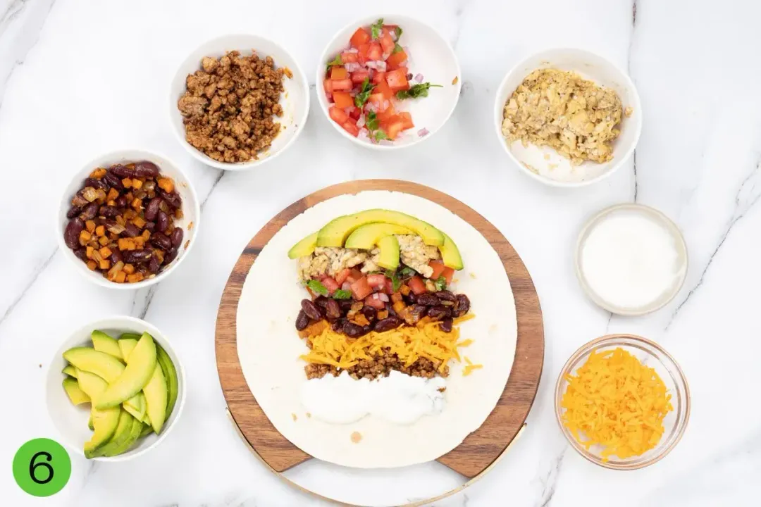 A burrito station of a wooden board with corn tortillas and fillings on top, surrounded by colorful fillings in seven separate bowls