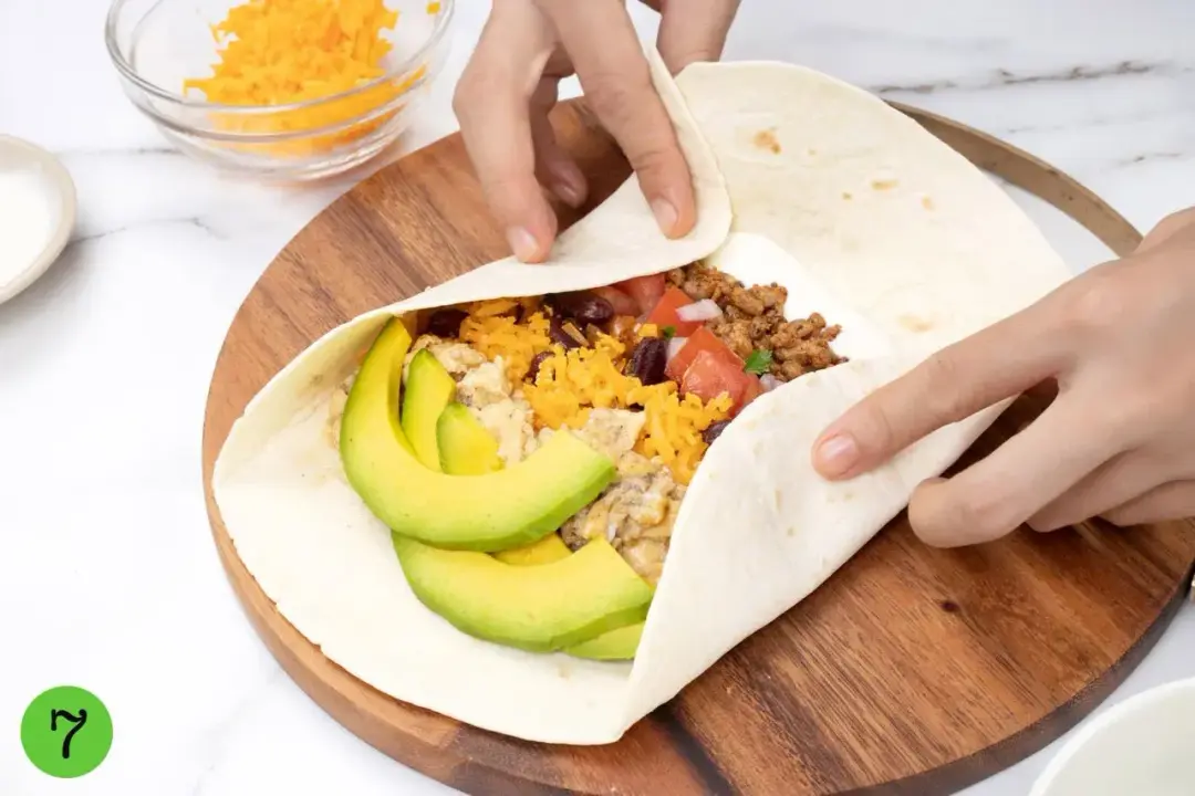 Two hands wrapping a flour tortilla and filling on top of a wooden board