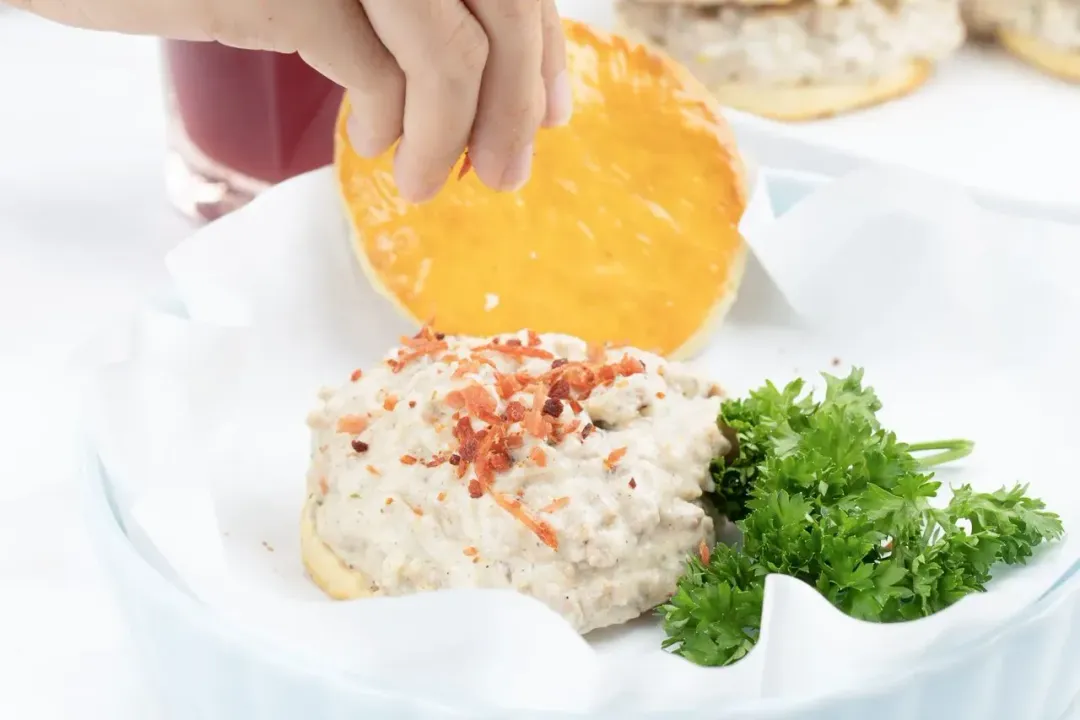 A hand placing a golden biscuit on a heap of white meat sauce and bacon, with parsley on the side