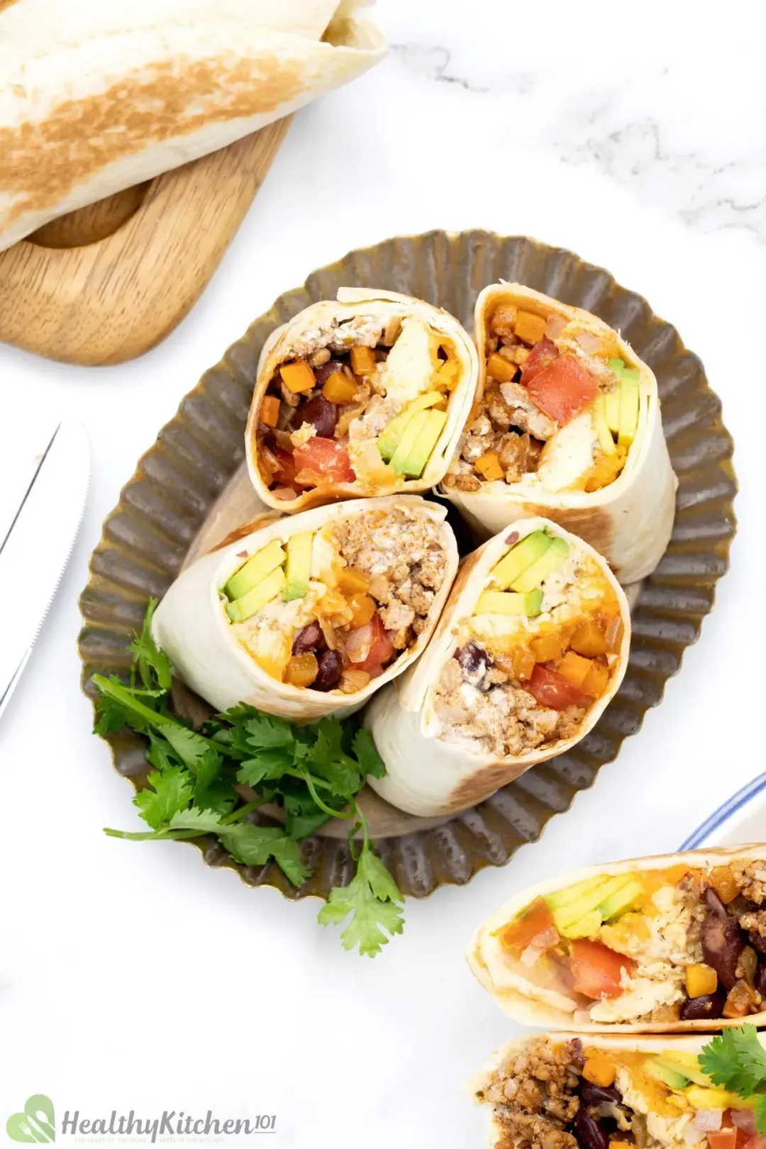 A plate of four burrito rolls with the cut side on top, showing colorful fillings, next to some cilantros and extra burrito rolls on the side