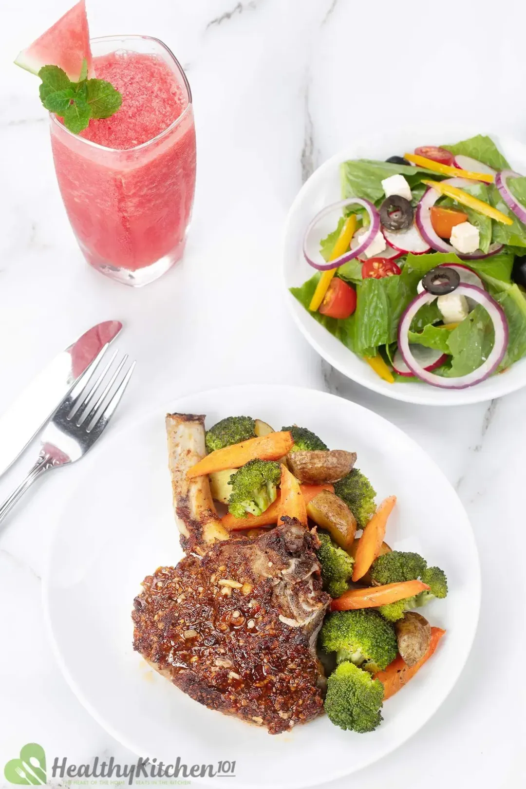 A portion of pork chop and roasted vegetables in a white plate, next to a greek salad and a watermelon juice