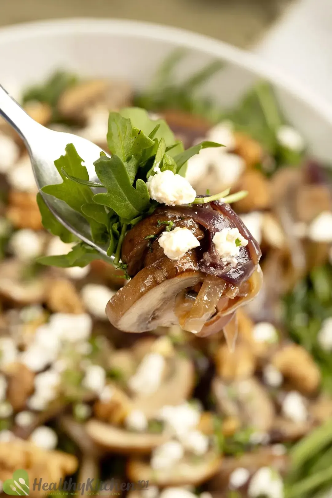 A close-up photo of a fork piercin through arugula mushroom salad with feta cheese, hovering over a bowl of salad