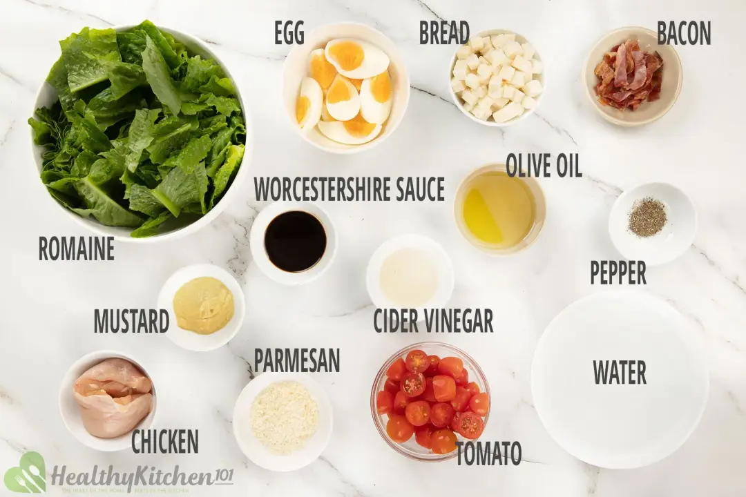 Ingredients for making Caesar salad including lettuce, croutons, parmesan cheese, boiled eggs, bacon crumbles, chicken, and dressing ingredients