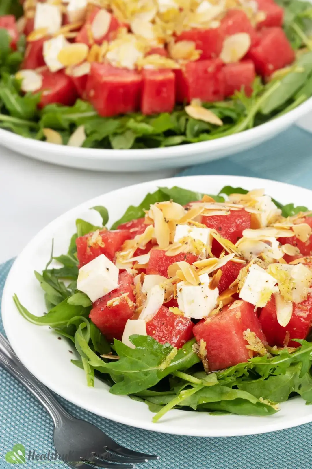 Two plates of watermelon arugula salad, topped with cubed feta cheese and shaved almonds