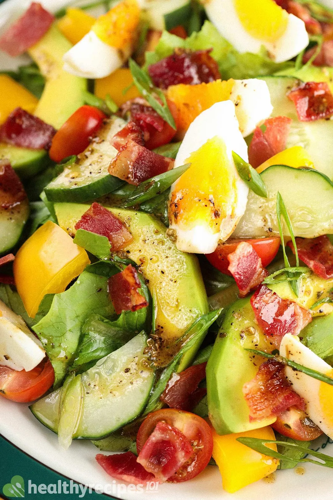 A close-up shot of chopped salad filled with sliced avocados, boiled eggs, halved cherry tomatoes, red and yellow bell peppers, and chopped scallions