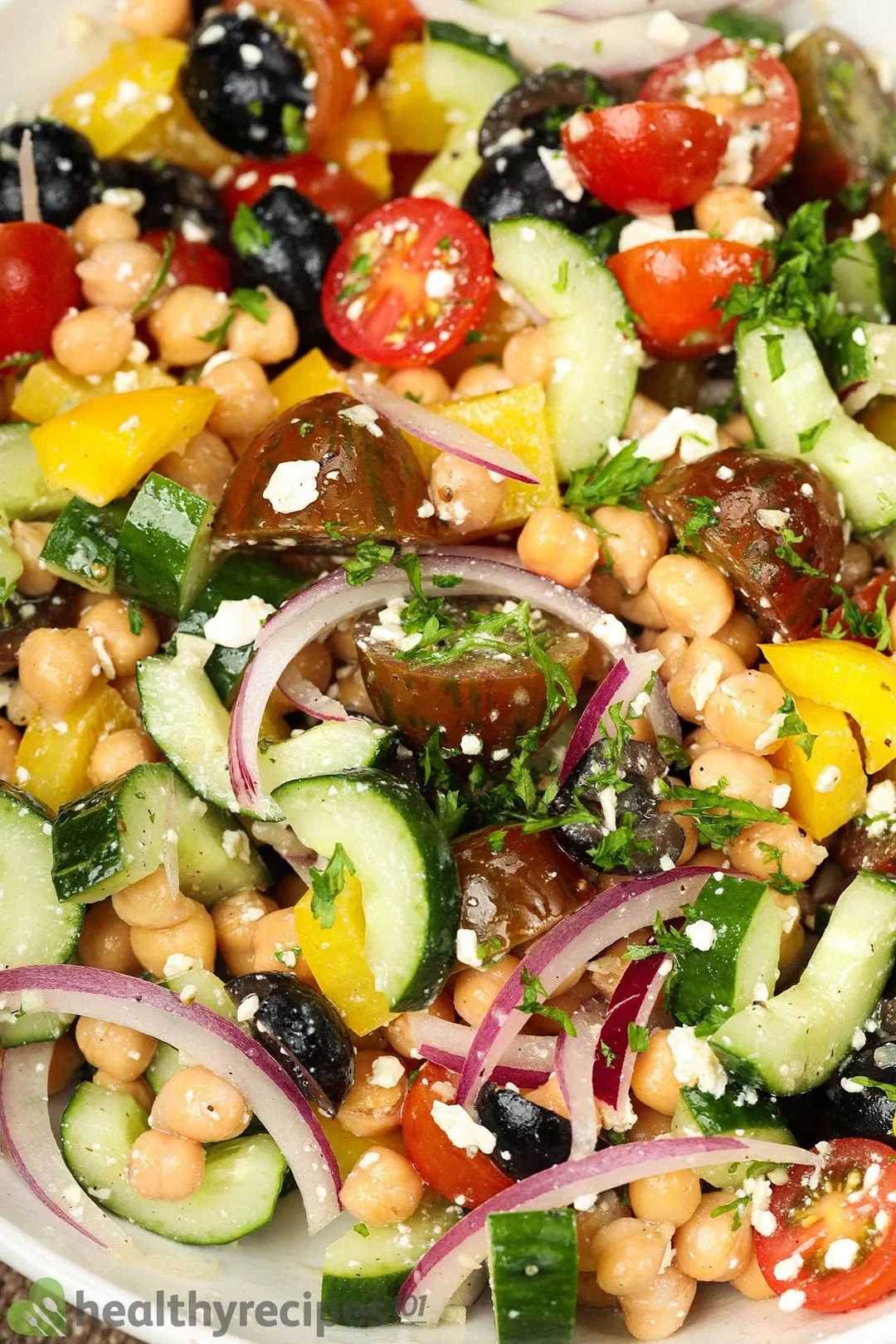 A close-up shot of a Greek chickpea salad, showcasing pieces of red onion, chickpeas, cherry tomatoes, yellow bell peppers, and black olives.